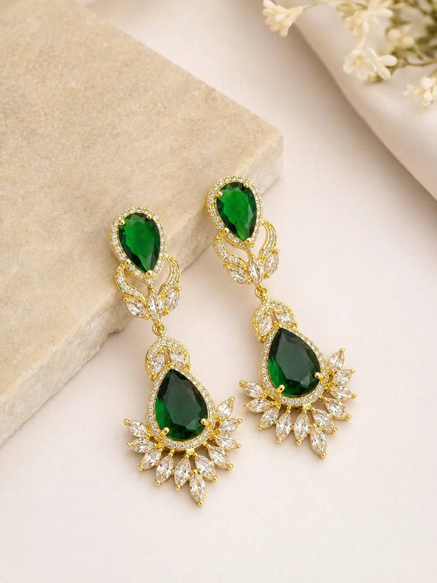 Pair of gold-plated emerald green CZ drop earrings laid flat on a beige stone surface
