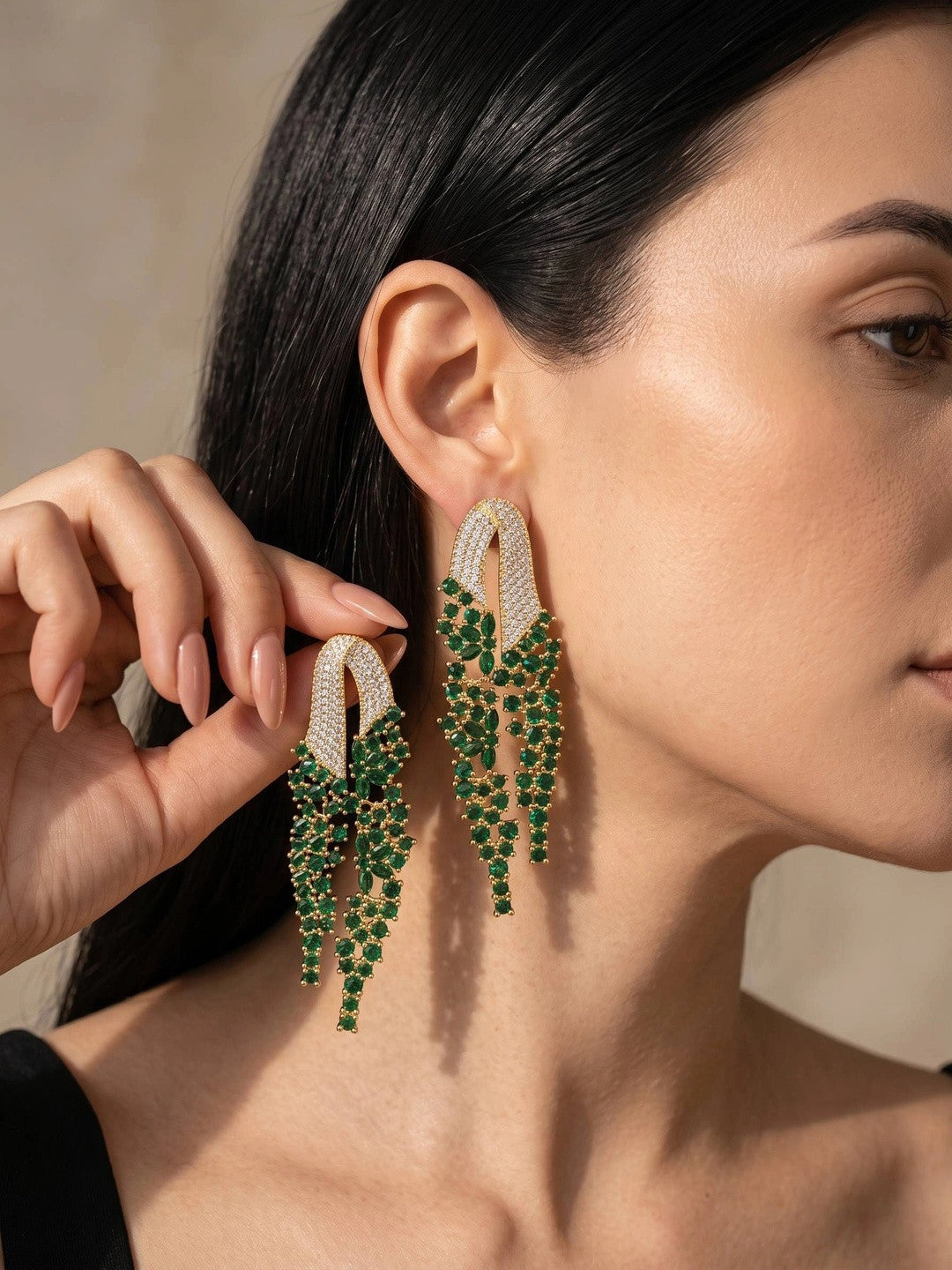 Woman wearing gold-plated emerald green CZ tassel drop earrings, holding one up to show the cascading waterfall design