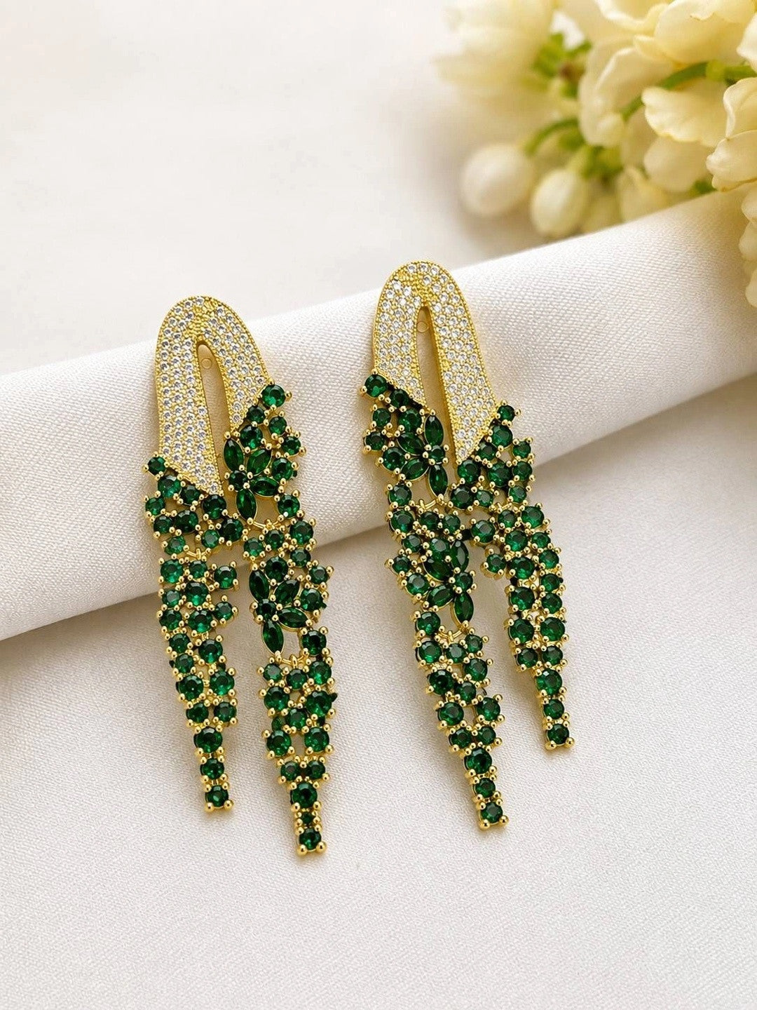 Pair of emerald green CZ tassel drop earrings laid flat on white fabric beside cream flowers, showing full front detail