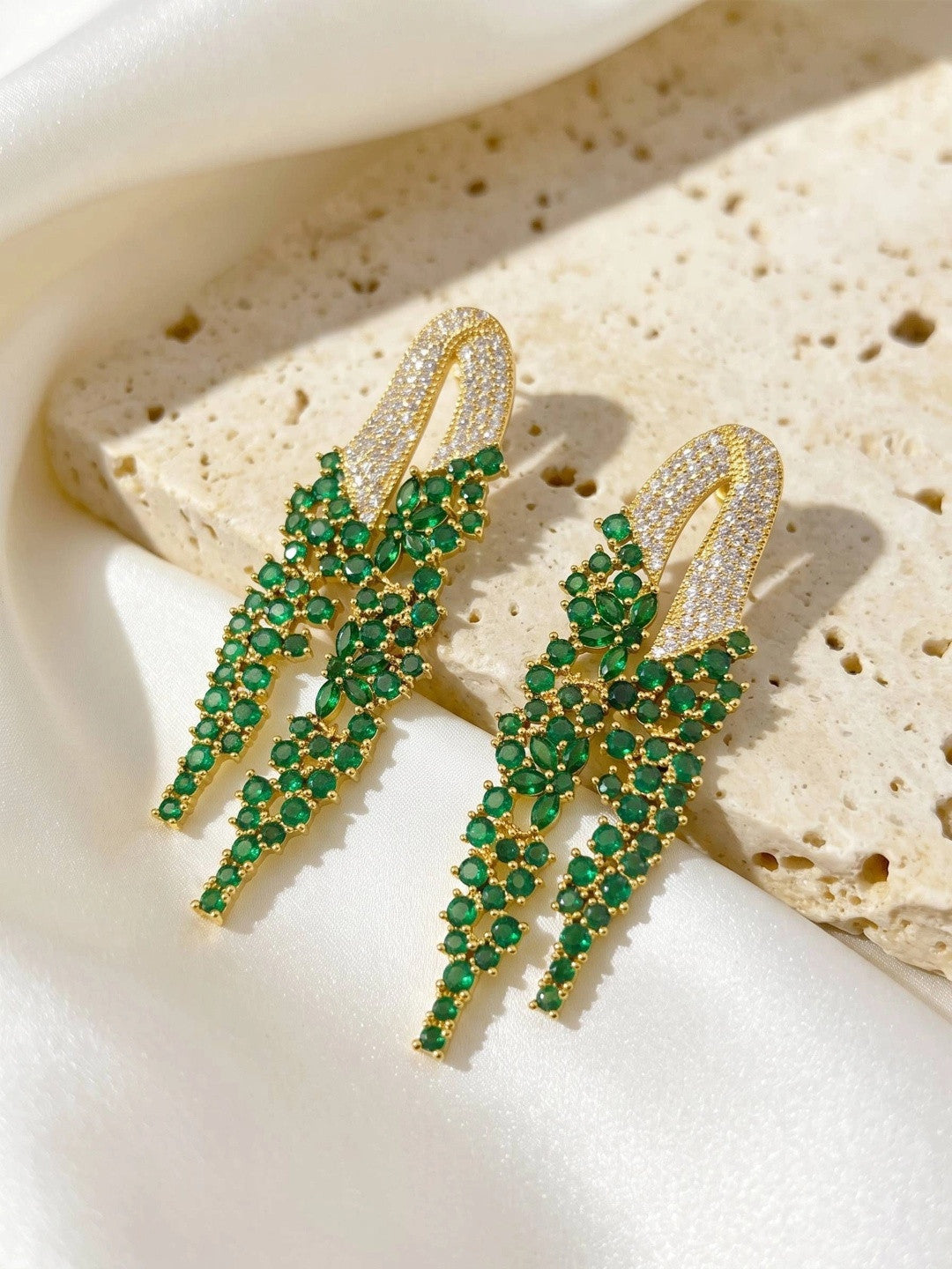 Two gold-plated emerald CZ tassel earrings resting on a textured stone surface in bright natural light