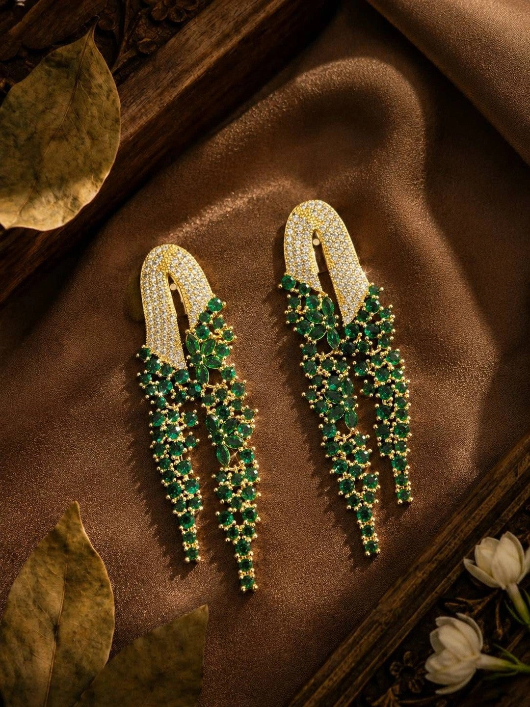 Emerald green CZ waterfall drop earrings displayed on brown silk fabric with dried leaves and white jasmine flowers