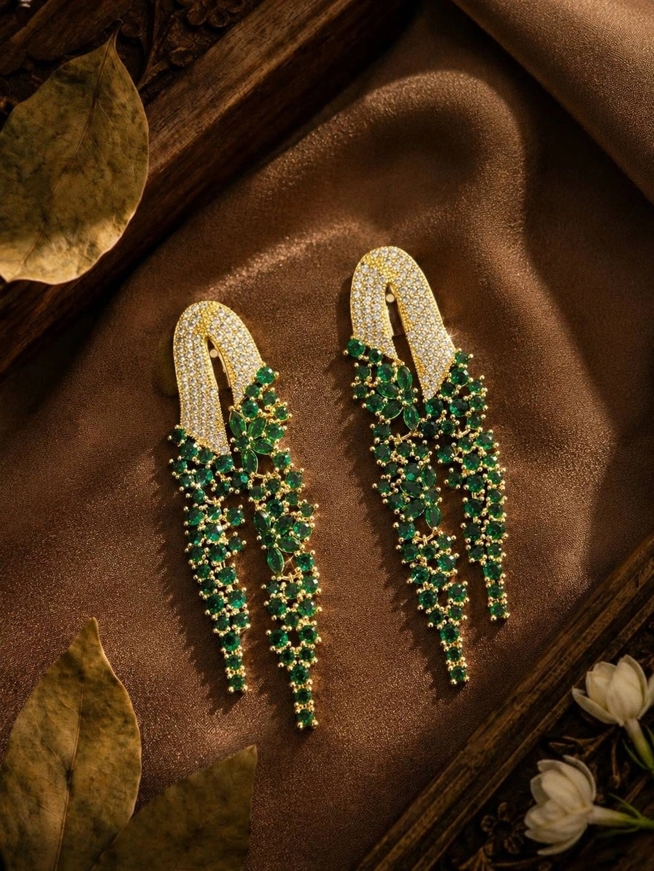 Emerald green CZ waterfall drop earrings displayed on brown silk fabric with dried leaves and white jasmine flowers