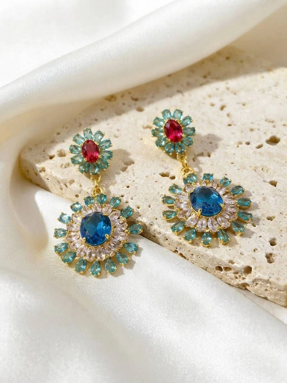 Two gold-plated teal and blue crystal drop earrings laid flat on a white stone surface