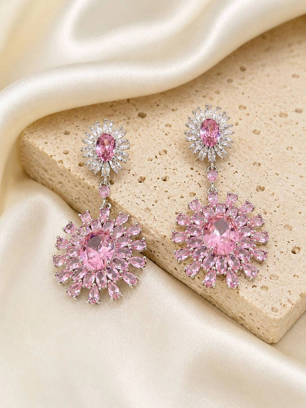Pair of pink crystal sunburst drop earrings laid on a beige stone surface with cream satin fabric