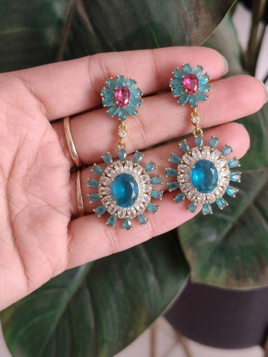 Both blue zircon drop earrings held in an open palm against green foliage background