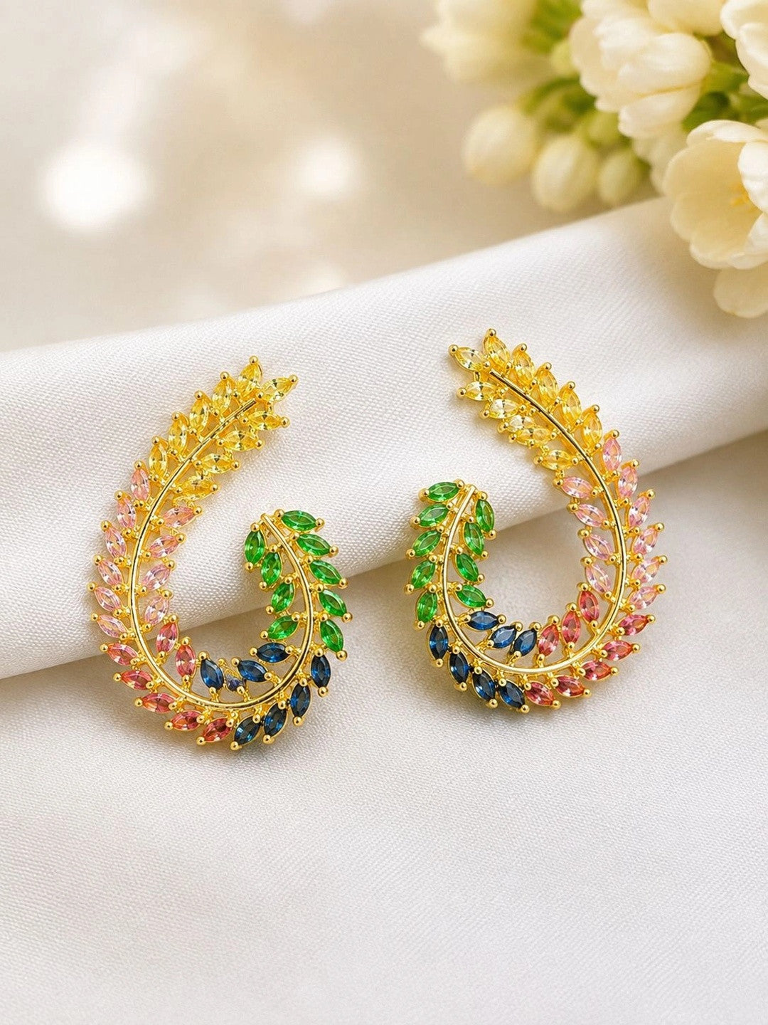 Pair of gold-plated multicolour CZ leaf half-hoop earrings laid flat on white fabric beside cream flowers