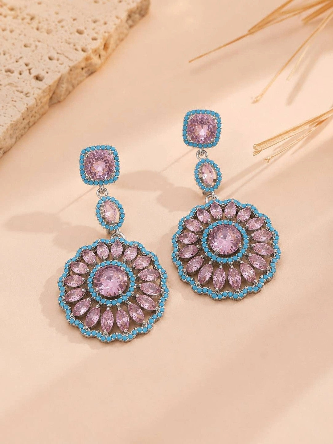 Pink and blue zirconia circular drop earrings displayed on a beige surface with dried grass stems