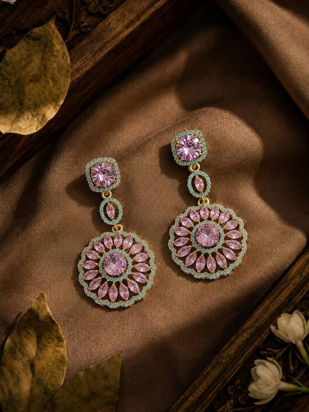 Two gold-plated circular drop earrings with pink stones and turquoise border displayed on golden fabric with dried leaves