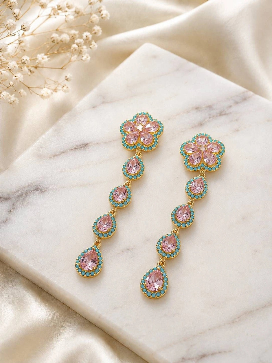 Two gold-tone floral drop earrings with pink teardrop stones and teal pavé laid on white marble