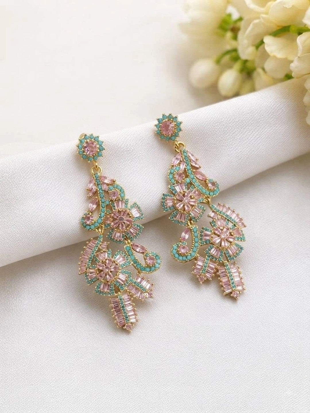Two gold-plated pink and teal crystal chandelier earrings laid flat on white fabric with flowers