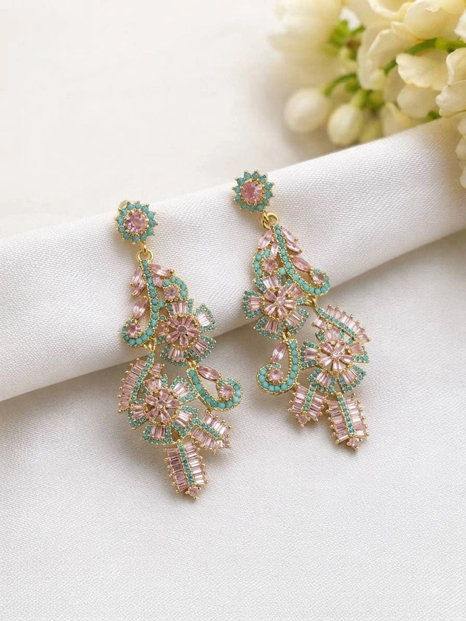 Two gold-plated pink and teal crystal chandelier earrings laid flat on white fabric with flowers