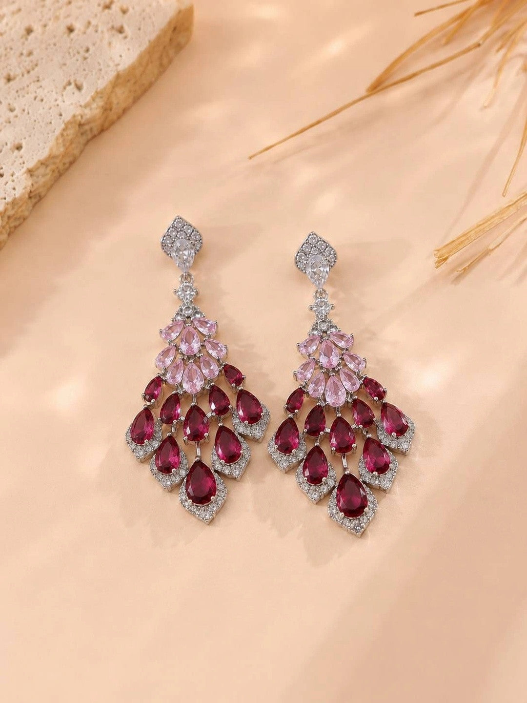 Ruby pink CZ drop earrings on a peach surface with dried grass stems in the background
