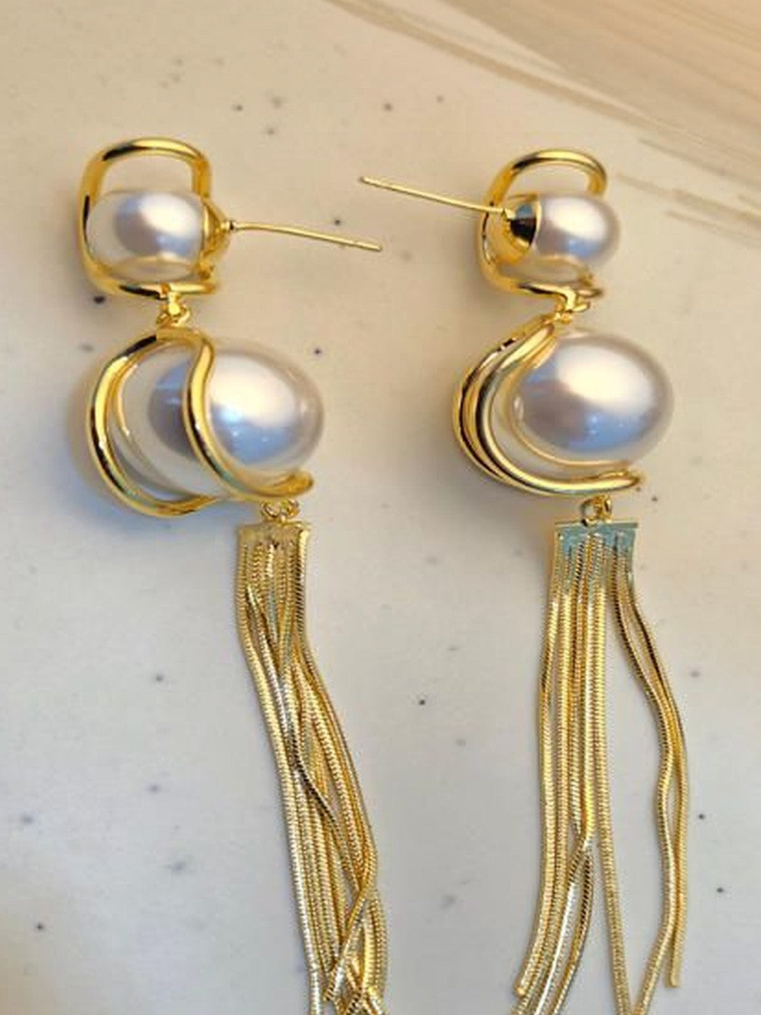 Pair of gold-plated faux pearl tassel earrings laid flat on a white marble surface showing post studs