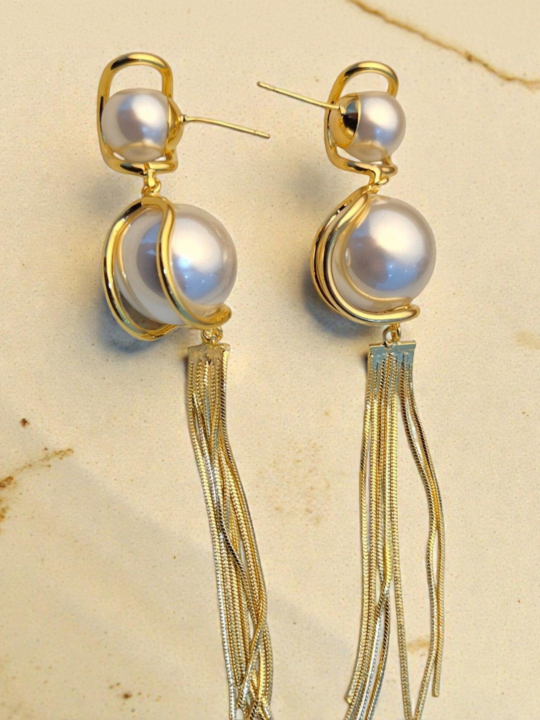 Gold swirl-set faux pearl drop earrings with long multi-strand snake-chain tassels on a cream stone background