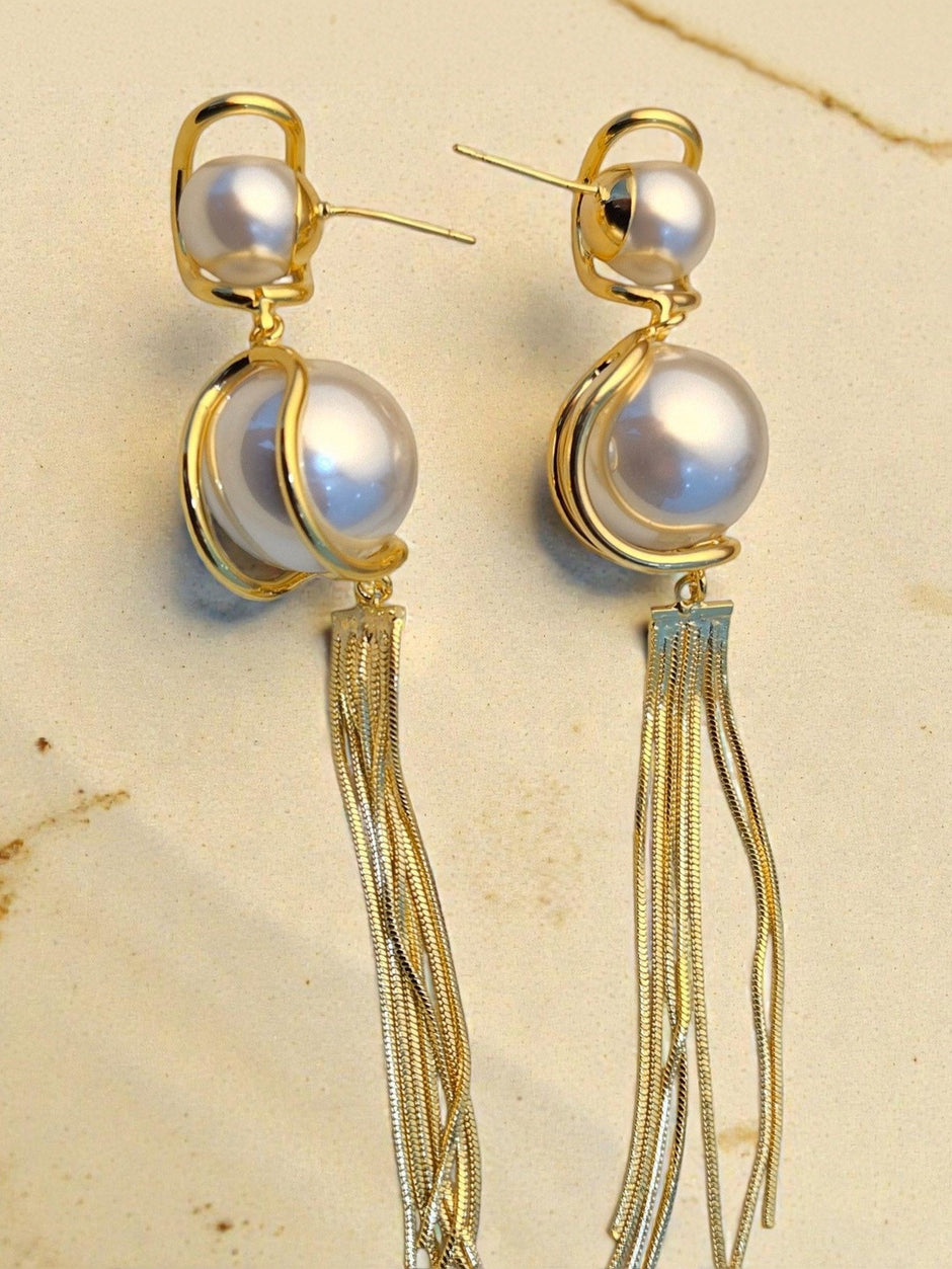 Gold swirl-set faux pearl drop earrings with long multi-strand snake-chain tassels on a cream stone background