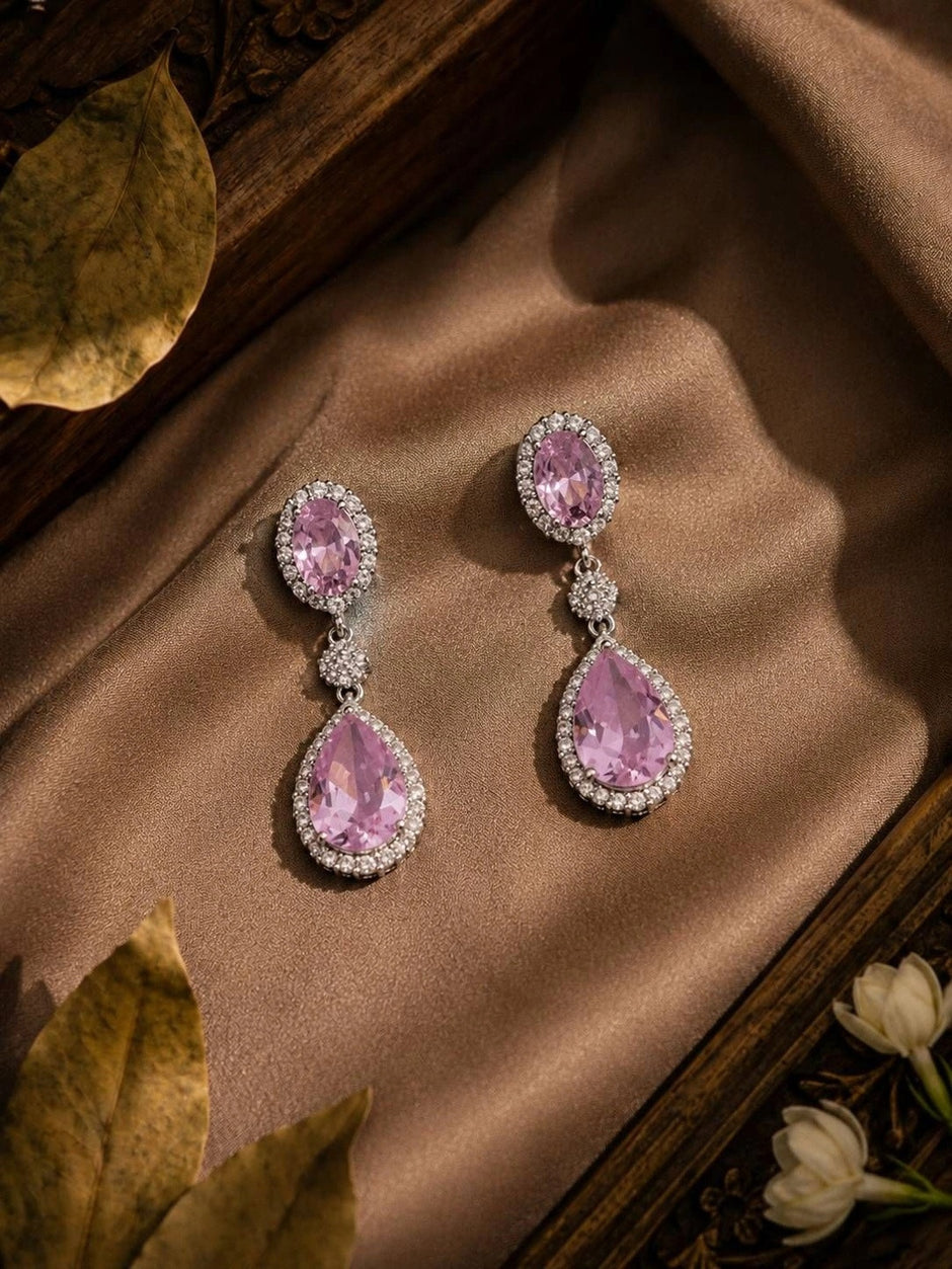 Pair of pink CZ teardrop drop earrings laid on bronze satin fabric with dried leaves and white flowers