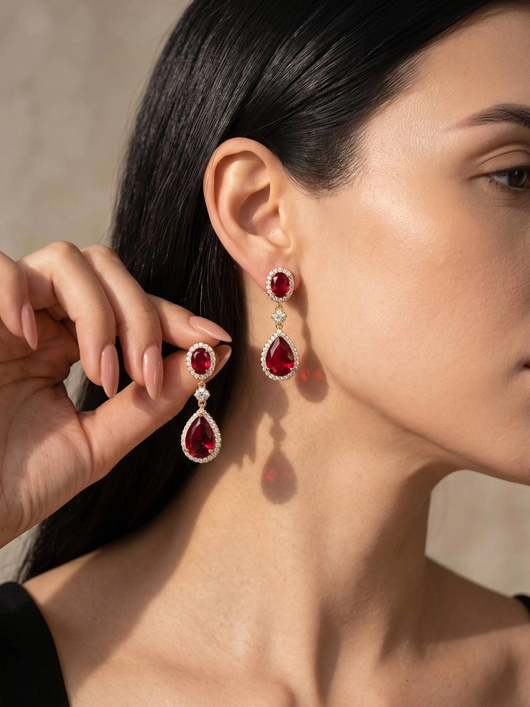 Woman wearing ruby red gold-plated teardrop drop earrings, holding one up to show detail