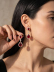 Woman wearing ruby red gold-plated teardrop drop earrings, holding one up to show detail
