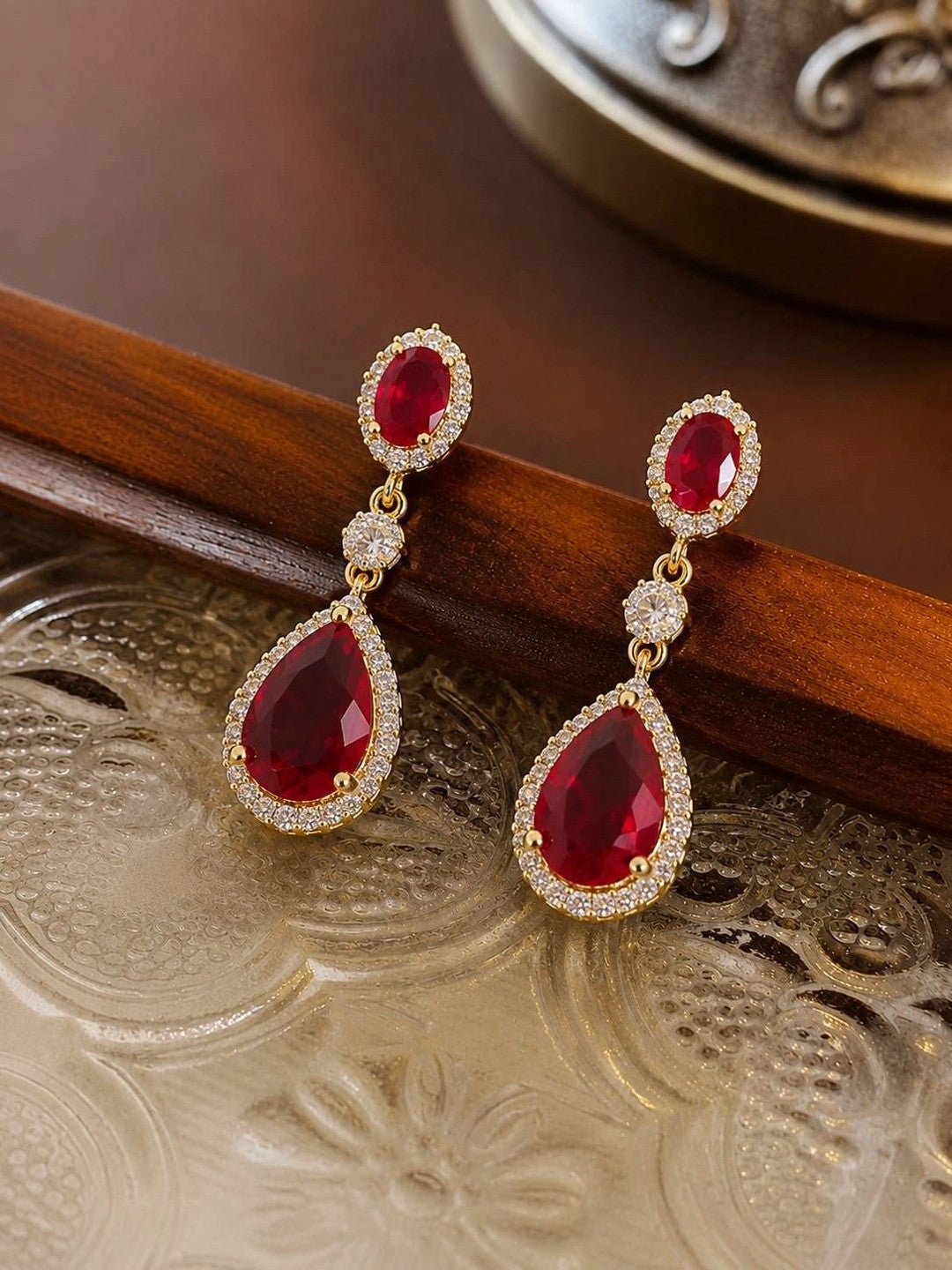 Pair of ruby red CZ teardrop drop earrings resting on a decorative gold and wood tray
