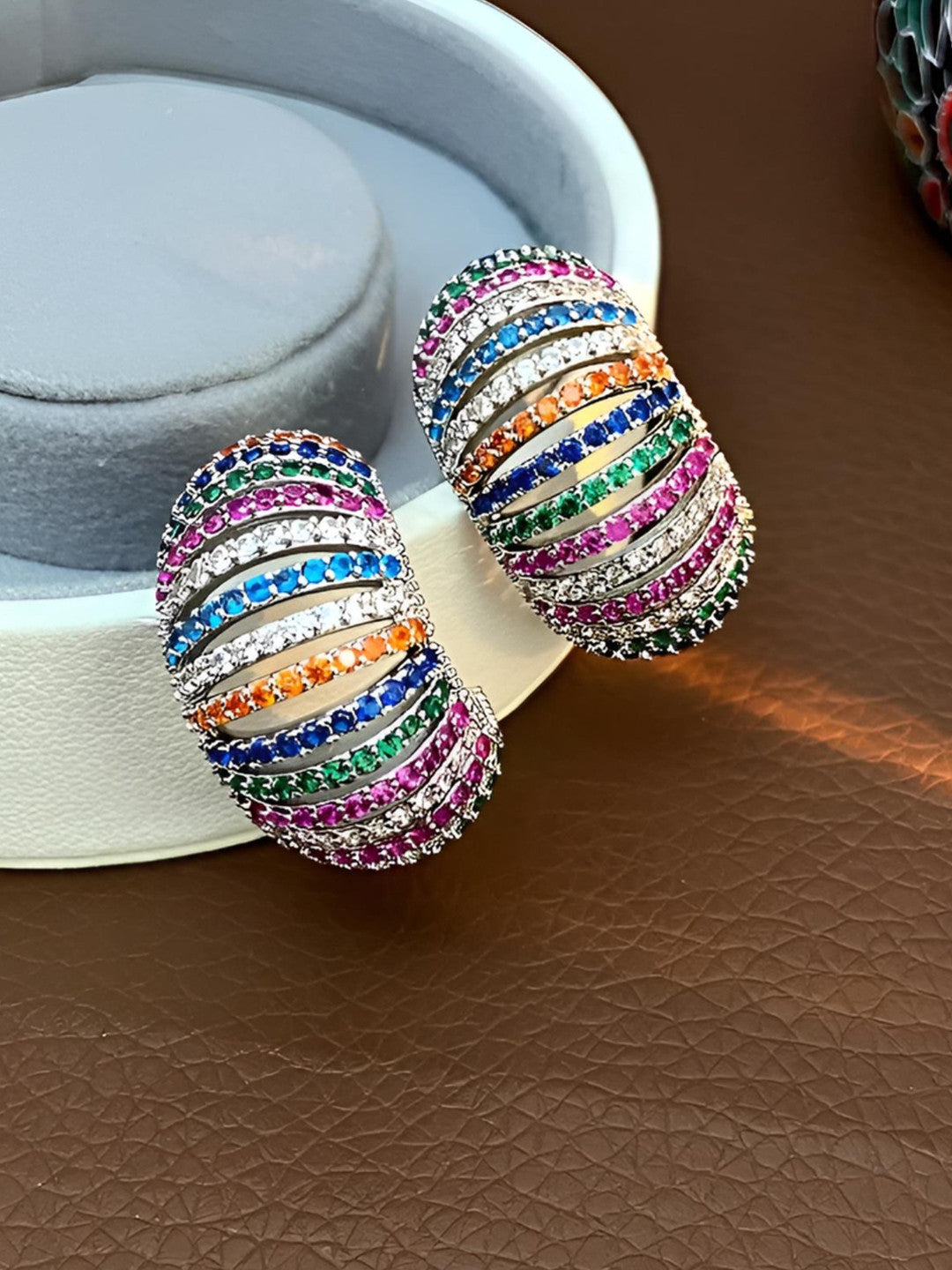 Pair of multicolour zirconia chunky hoops resting against a white jewellery box on a brown leather surface
