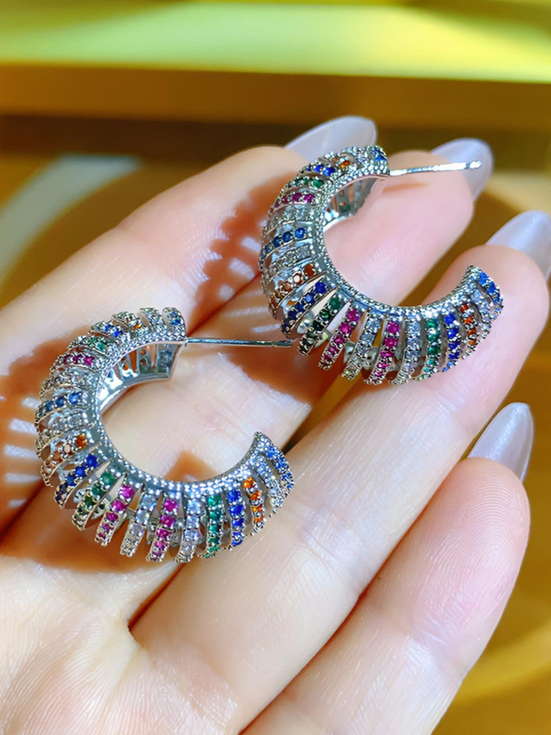 Close-up side profile of two rainbow zirconia C-hoop earrings held in hand showing full arc and stone rows