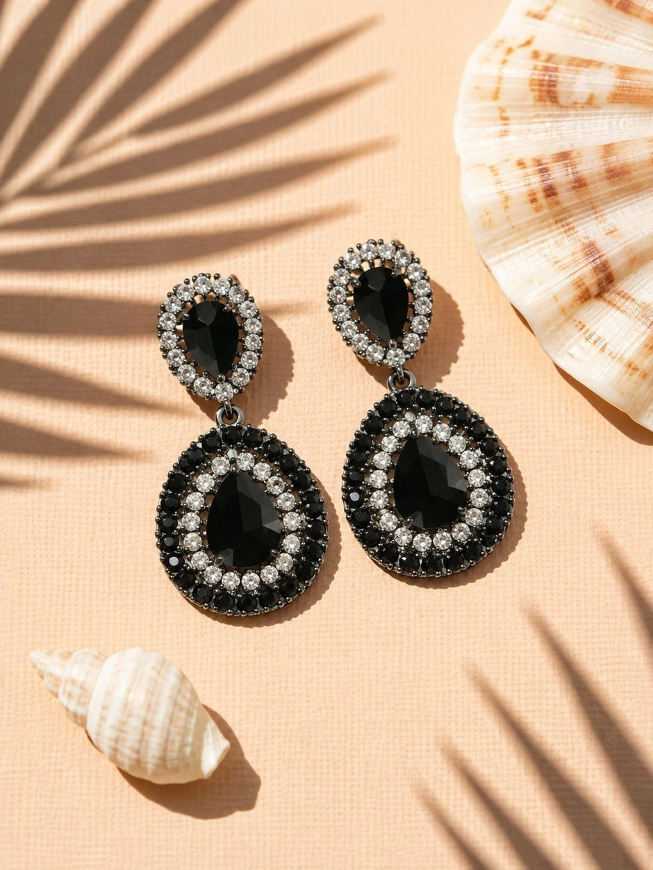Pair of black and clear crystal teardrop drop earrings styled with seashells on peach background