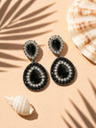 Pair of black and clear crystal teardrop drop earrings styled with seashells on peach background