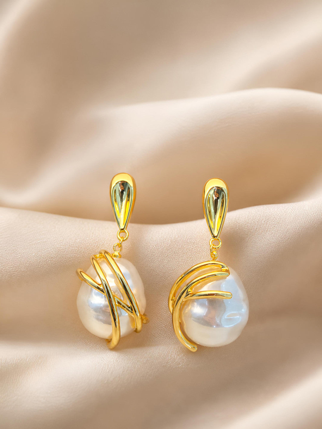 Pair of gold-plated baroque pearl drop earrings laid on cream satin fabric, showing criss-cross metal detail