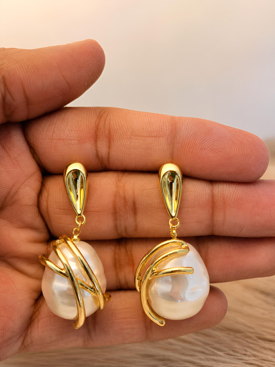 Close-up of two gold-plated baroque pearl drop earrings held in an open palm, showing scale and finish detail
