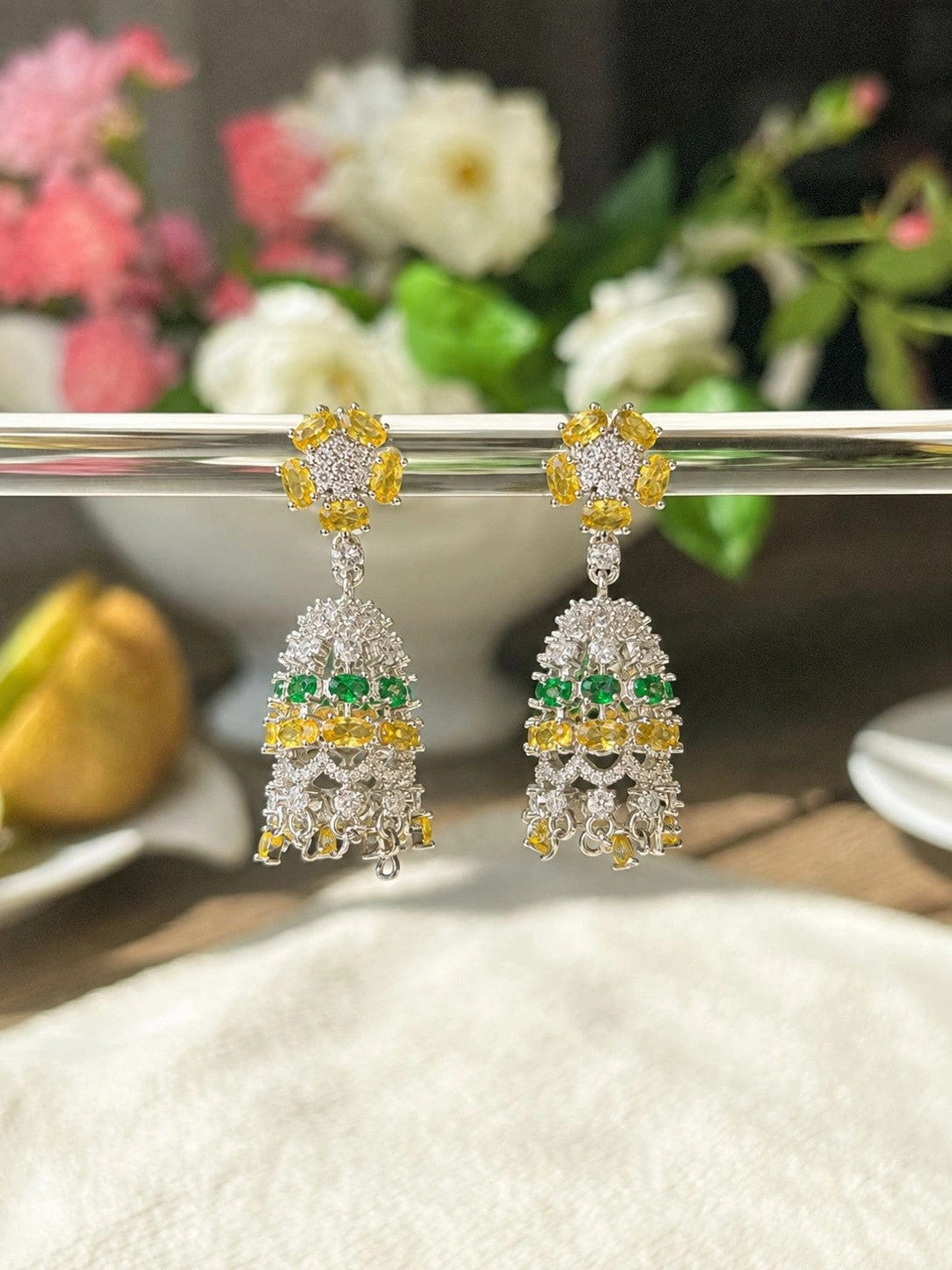 Pair of silver CZ chandelier earrings hanging from metal bar with floral backdrop in soft light