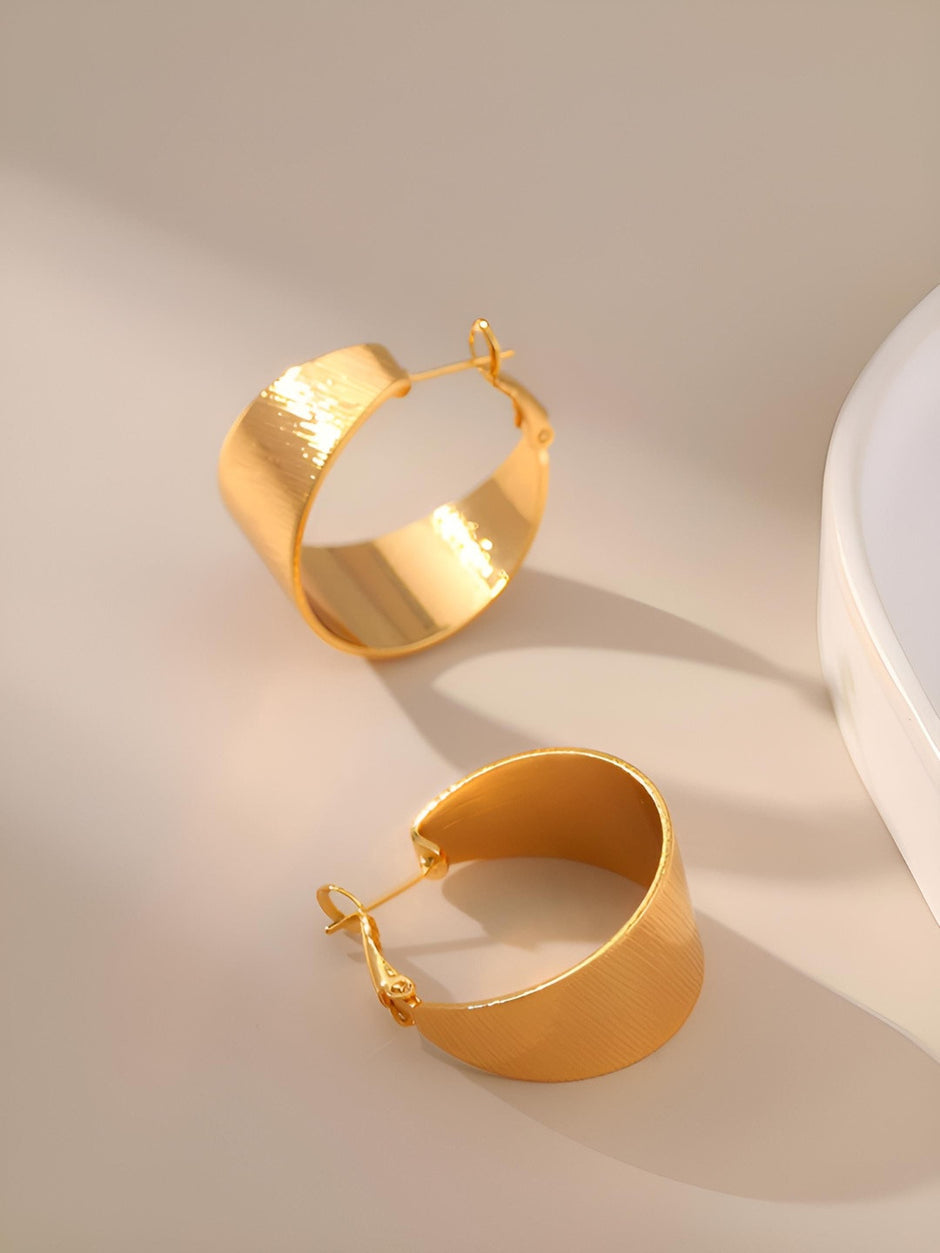Two gold-plated wide band hoop earrings lying flat on a beige surface showing inner polished finish