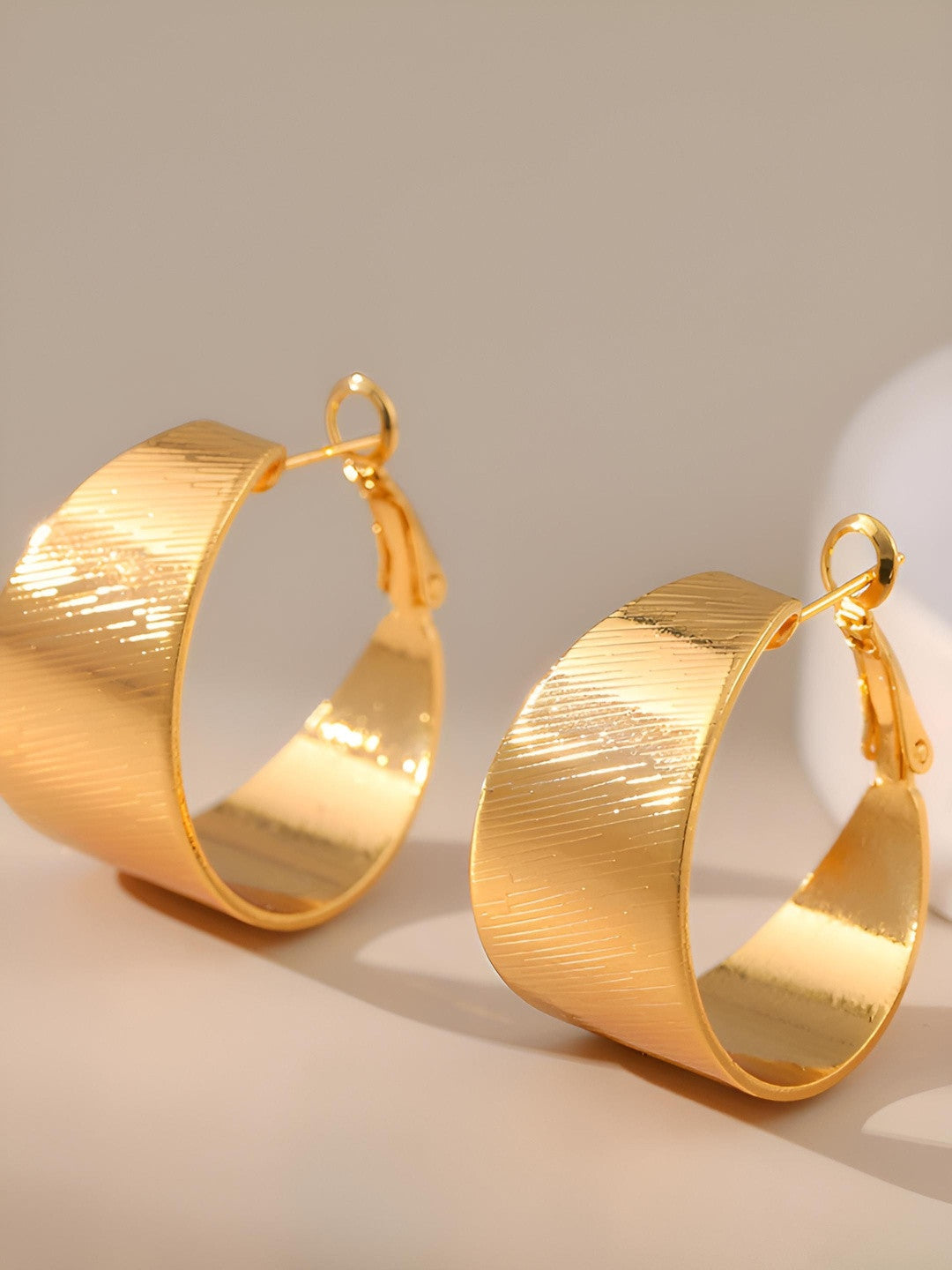 Pair of gold-plated textured wide hoop earrings standing upright on a light grey surface