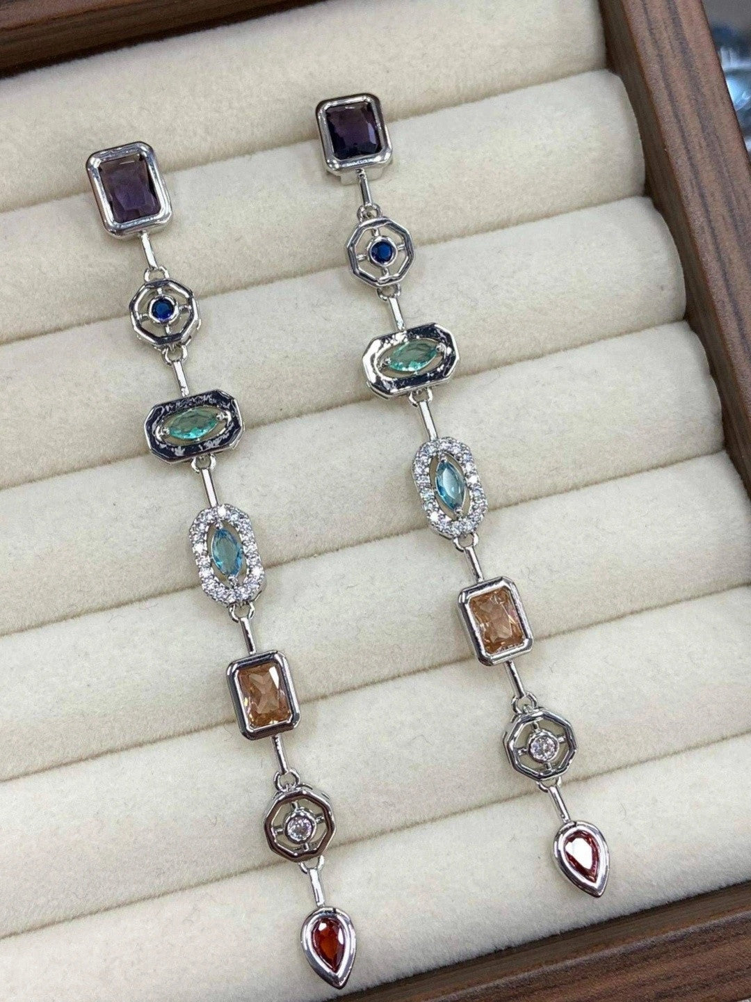 Pair of silver-tone geometric drop earrings laid flat in a cream jewellery box, displaying all six coloured stone stations from above