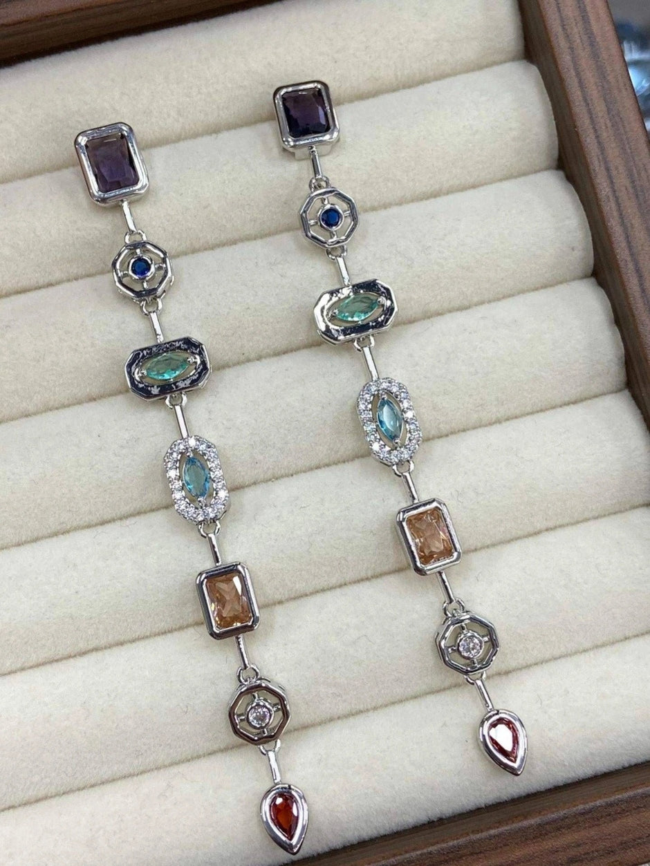 Pair of silver-tone geometric drop earrings laid flat in a cream jewellery box, displaying all six coloured stone stations from above