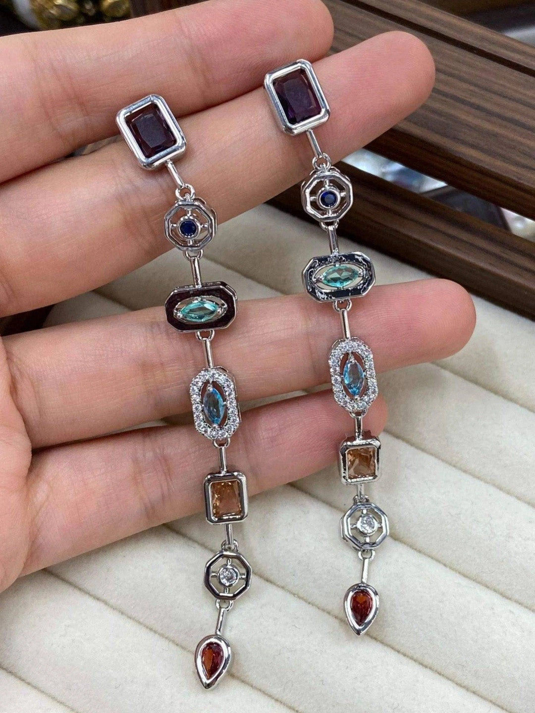 Two silver-tone multi-stone dangler earrings held in a hand against a display tray, showing full length and stone arrangement