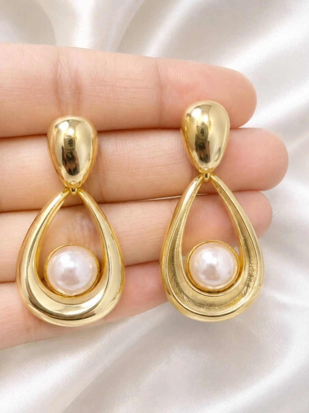 Pair of gold-plated pearl drop earrings held in an open palm against white satin fabric