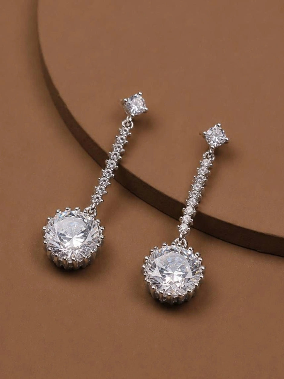 Silver-tone CZ halo drop earring pair laid on a warm brown display surface at an angle