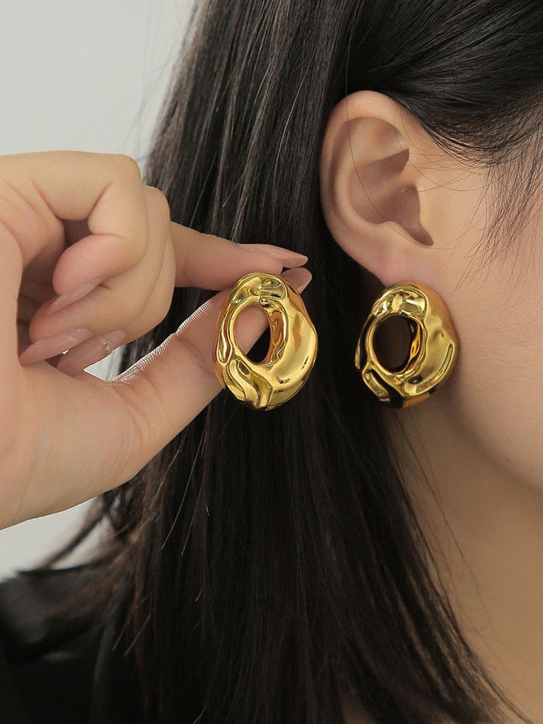 Woman wearing gold hollow oval stud earrings held up to ear showing scale and fit