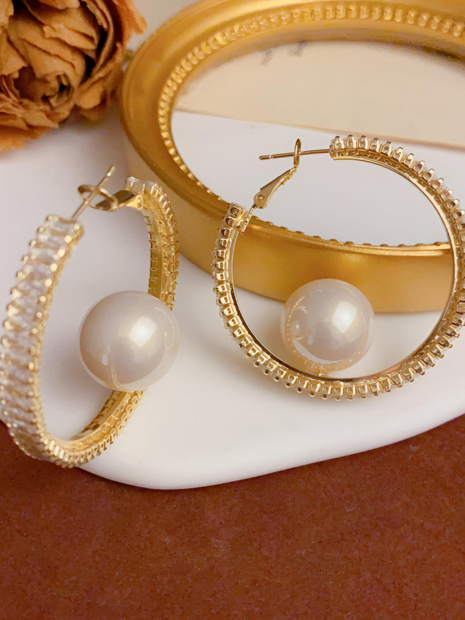 Two gold-plated pearl hoop earrings displayed against a gold frame and white ceramic surface