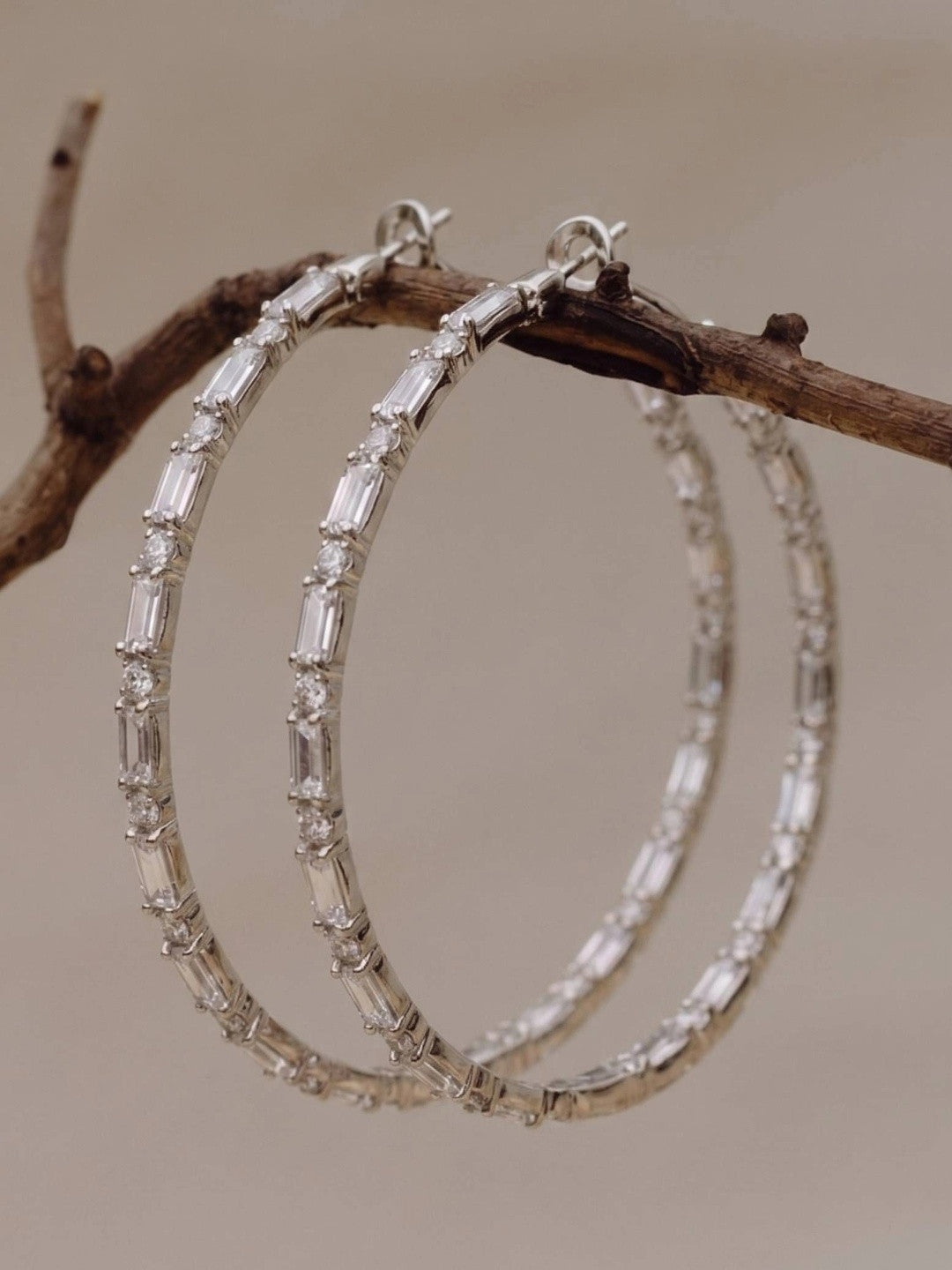 Pair of silver CZ hoop earrings resting on a wooden branch, showing baguette and round stone alternating pattern