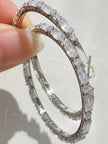 Close-up of two silver hoop earrings held between fingers, showing prong-set baguette cubic zirconia stones and latch-back closure