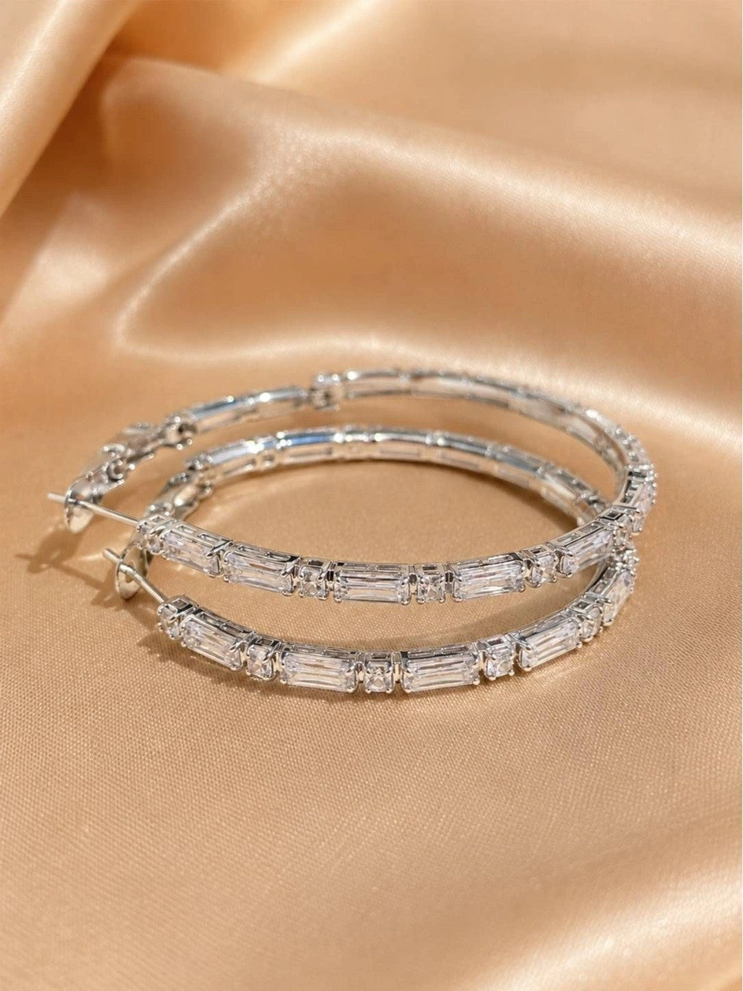 Pair of silver baguette CZ hoop earrings laid flat on golden satin fabric, post closures visible