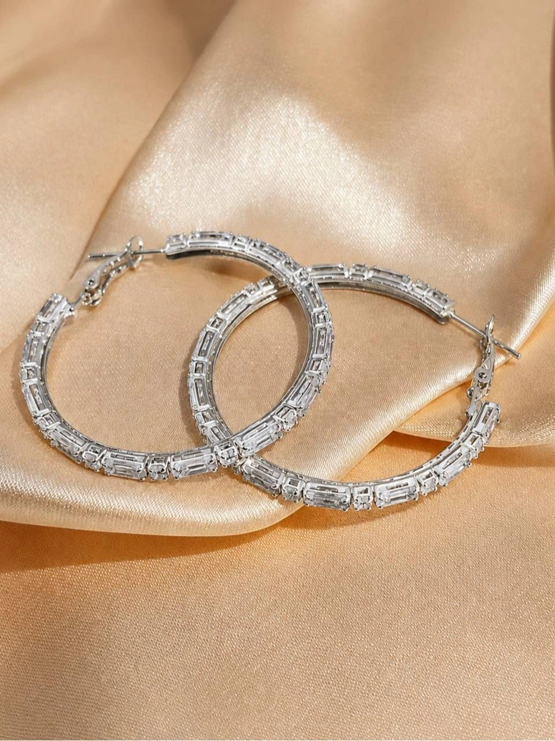Two silver cubic zirconia hoop earrings overlapping on champagne satin, full circular form and all-around stone setting visible