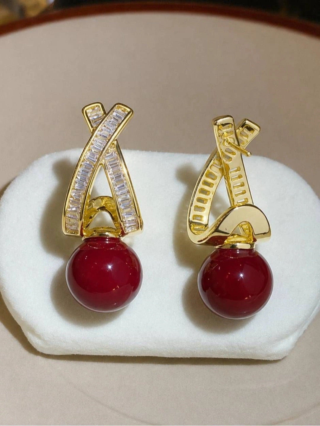 Front and back view of gold-plated X-drop earrings with red bead on white jewellery cushion