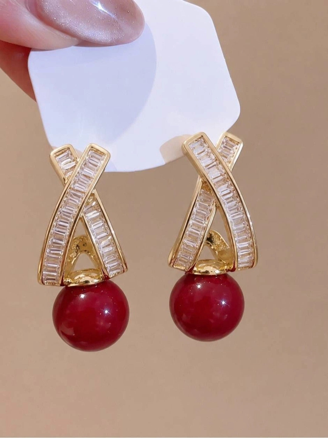 Pair of gold crystal criss-cross drop earrings with crimson bead held against beige background