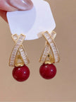 Pair of gold crystal criss-cross drop earrings with crimson bead held against beige background