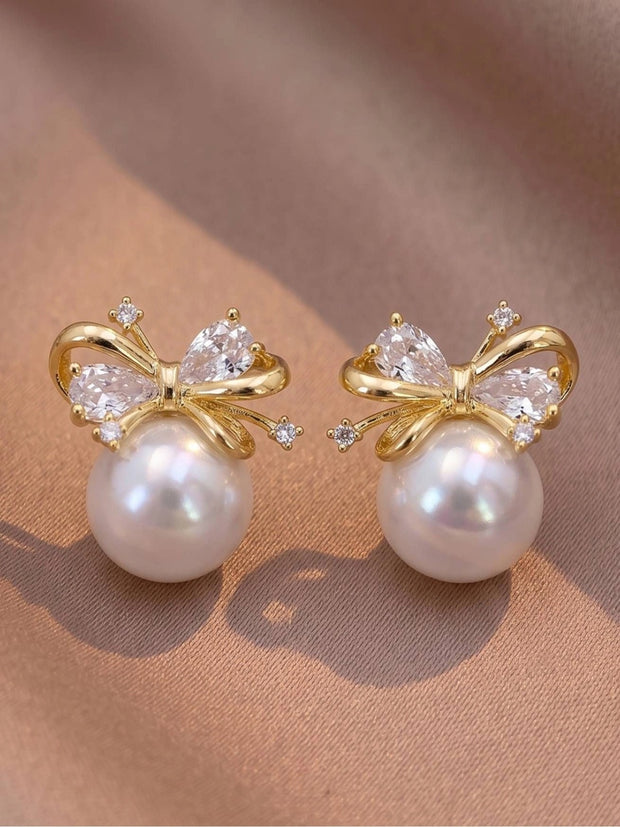 Pair of gold-plated bow stud earrings with faux pearl and cubic zirconia stones on a pink fabric surface
