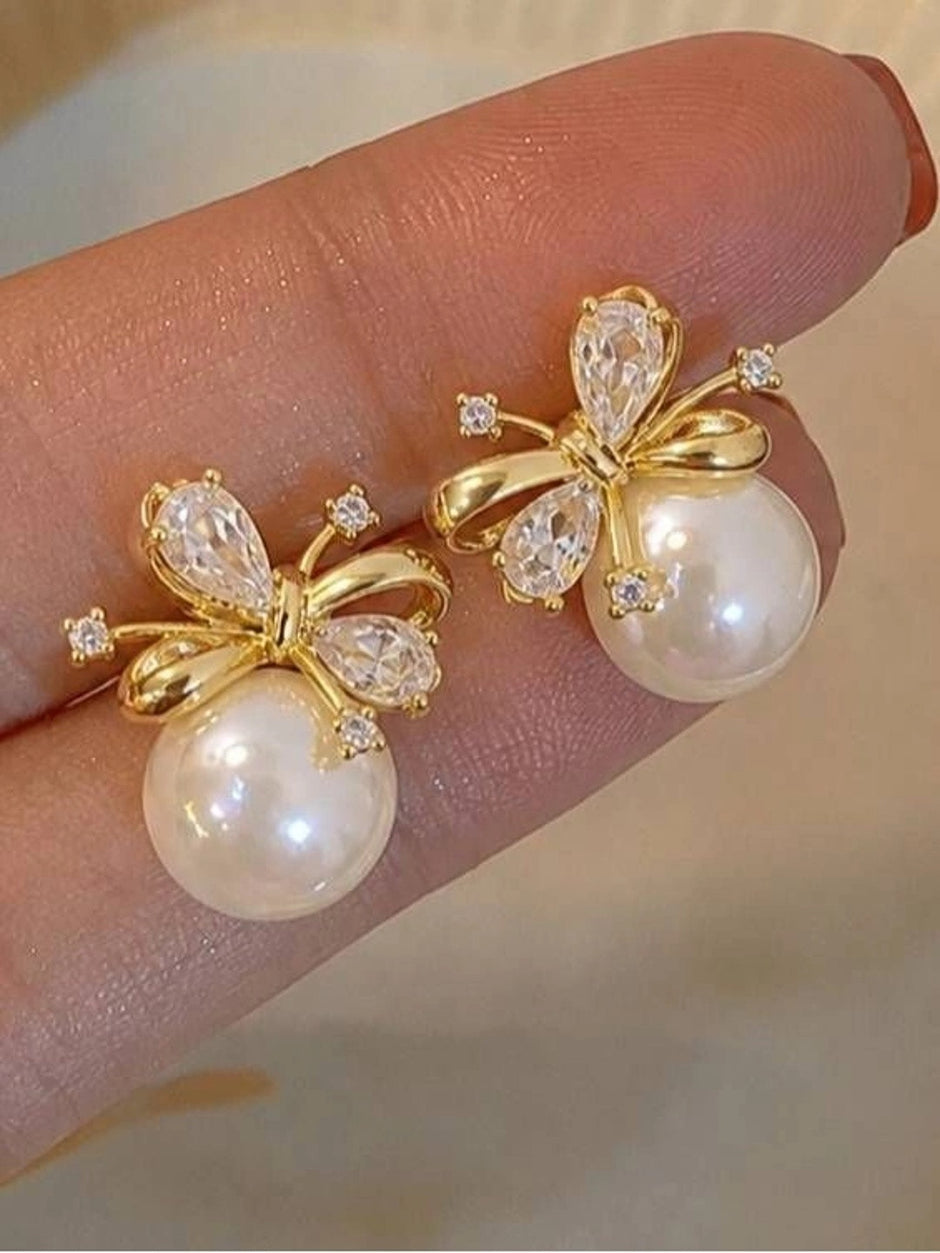 Two gold-plated pearl bow stud earrings resting on a model's fingertips showing CZ detail