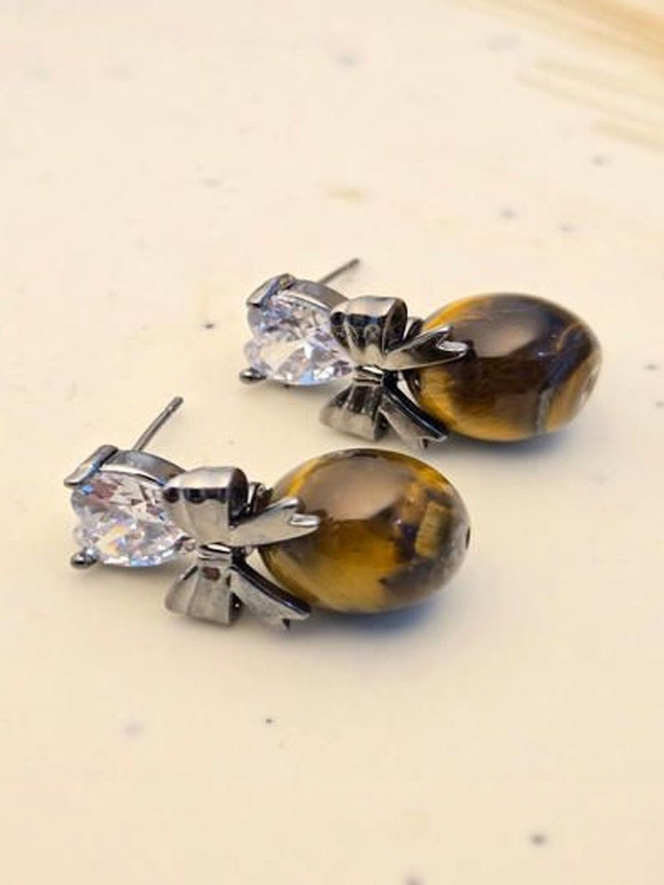 Pair of oxidised silver bow drop earrings with heart cubic zirconia and oval tiger eye beads on cream surface
