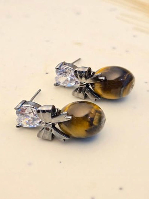 Pair of oxidised silver bow drop earrings with heart cubic zirconia and oval tiger eye beads on cream surface